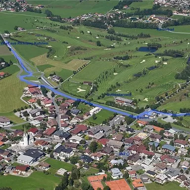 Eine Luftaufnahme eines kleinen Dorfes mit saftig grünen Feldern und einem Golfplatz. Im Vordergrund sind Wohnhäuser und eine Kirche sichtbar.