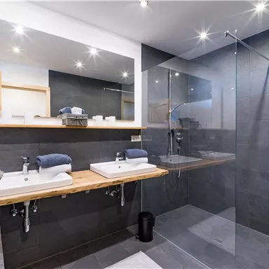 A modern bathroom with two sinks and a glass shower. The walls are made of dark stone and there is ample lighting.