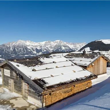 A snowy mountain landscape with two old wooden chalets. The clear blue sky and the surrounding mountains create an idyllic atmosphere.