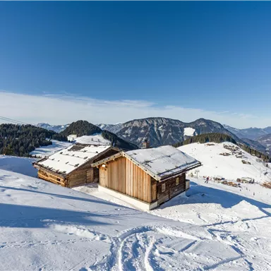 A snowy mountain landscape with traditional cottages and a blue sky. In the distance, the snow-covered mountains are visible.