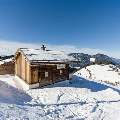 A cozy wooden cottage in the snow. In the background, mountains and a cable car can be seen.