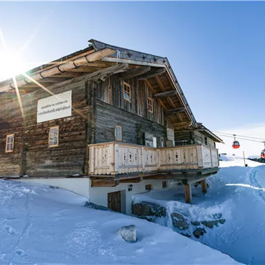 A cozy wooden house in a snowy landscape. In the background, ski lifts can be seen, and the sun is shining brightly in the sky.