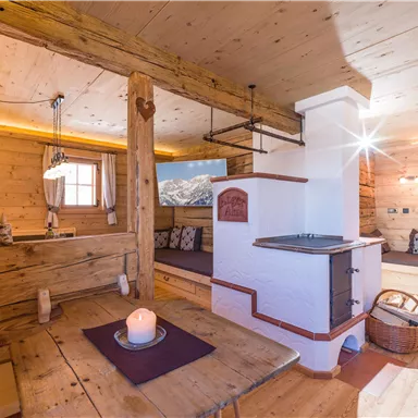 A cozy wooden cabin with a warm atmosphere. The space is furnished with a tiled stove, a dining table, and rustic furniture.