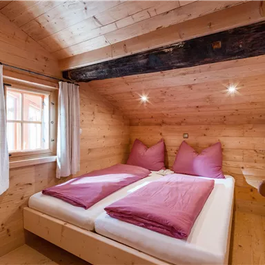 A cozy bedroom with wood paneling and a double bed. The bedding is in shades of purple and there are large windows for natural light.