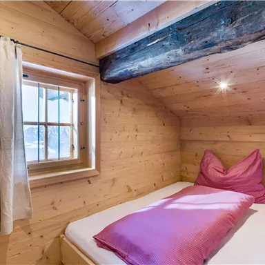 A cozy wooden bedroom with a window and simple pink bedding. The wooden paneling creates a warm atmosphere.