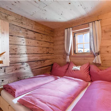 A cozy bedroom with wooden paneling and pink cushions. The window lets warm light in, creating an inviting atmosphere.