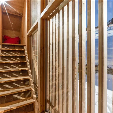 A bright wooden staircase in a modern building. Wooden slats are on the walls and a red cushion is in the background.