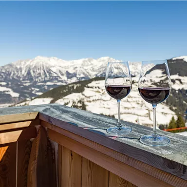 Two glasses of red wine on a wooden terrace with snow-covered mountains in the background. The sky is clear and blue.