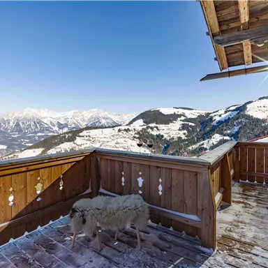 A balcony with a wooden floor and a beautiful view of snow-covered mountains. A sheep is standing on the balcony, enjoying the winter landscape.