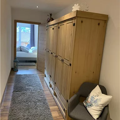 A narrow hallway with a wooden cabinet and a cozy chair. In the background, there is a window that lets light into the room.