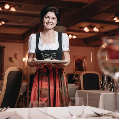 Eine Frau in traditioneller Tracht steht in einem eleganten Restaurant mit einem Teller in der Hand. Im Hintergrund sind gedeckte Tische und warme Lichtquellen zu sehen.