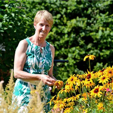 Eine lächelnde Frau steht in einem bunten Garten voller gelber Blumen. Die Sonne scheint und die Umgebung wirkt freundlich und einladend.