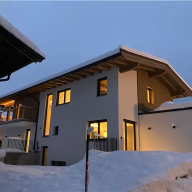 Ein modernes Haus im Schnee mit einer beleuchteten Fassade. Die Umgebung ist winterlich und ruhig.