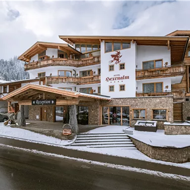 Ein schönes Hotel mit traditioneller Holzarchitektur in einer winterlichen Umgebung. Die Fassade ist teilweise mit Schnee bedeckt, umgeben von Bergen und Bäumen.