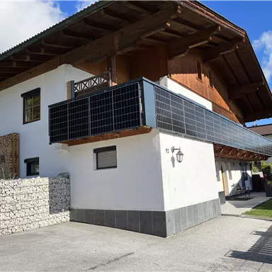 Ein modernes Haus mit einem Holzüberhang und einem Balkon. Die Fassade hat Solarpanels und es gibt eine Gartenansicht im Hintergrund.