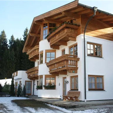 Ein schönes Haus im alpinen Stil mit Holzverkleidung und mehreren Balkonen. Umgeben von schneebedecktem Boden und Nadelbäumen.