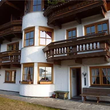 Ein charmantes, mehrstöckiges Haus im alpinen Stil mit Holzverzierungen und großen Fenstern. Es gibt Balkone und einen kleinen Gartenbereich vor dem Gebäude.
