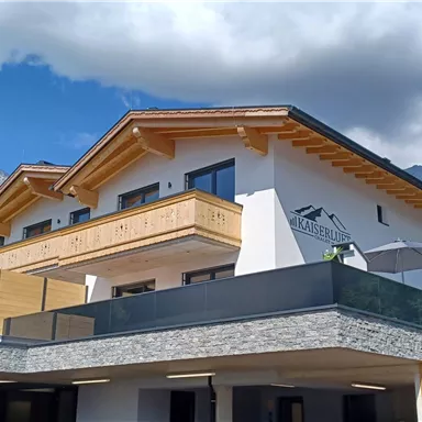 A modern building in the mountains with wooden cladding and large windows. The surroundings are green and surrounded by trees, under a clear sky.