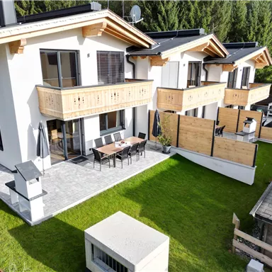 Two modern houses with wooden cladding in a green environment. On the terrace, there is a dining table and chairs.
