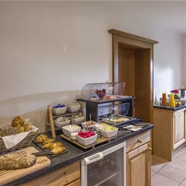 Eine gemütliche Küche mit einem reichhaltigen Frühstücksangebot. Es gibt frische Brötchen, Obst und Getränke auf einem dunklen Tisch.