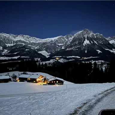 A snowy landscape at night with illuminated houses. In the background, impressive mountains rise under a clear starry sky.
