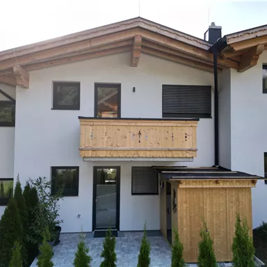 A modern facade of a house with a wooden balustrade and large windows. The entrance area is clearly designed and surrounded by small trees.