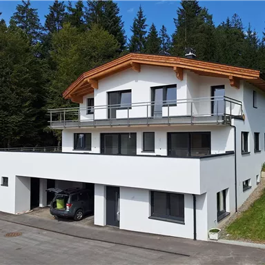 A modern, two-story house in a nature-friendly environment. It has a wooden roof overhang and appears bright and inviting.
