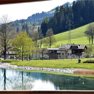 Eine idyllische Landschaft mit einem ruhigen See und grünen Wiesen. Im Hintergrund sind Häuser und verstreute Bäume vor einer sanften Hügelkulisse zu sehen.