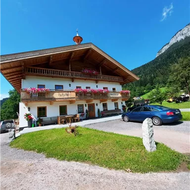 Ein traditionselles Haus in den Bergen mit einem schönen Balkon und Blumen. Ein blauer Wagen steht auf der Auffahrt vor dem Gebäude.