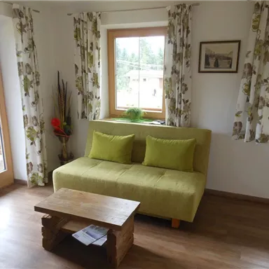 Ein gemütliches Wohnzimmer mit einem grünen Sofa und einem Holztisch. Große Fenster lassen viel Licht herein und es gibt dekorative Vorhänge.