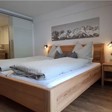 Ein modernes Schlafzimmer mit einem großen Holzbett und gemütlichen Kissen. An der Wand hängt ein Bild von Bergen, und es gibt eine helle, einladende Atmosphäre.