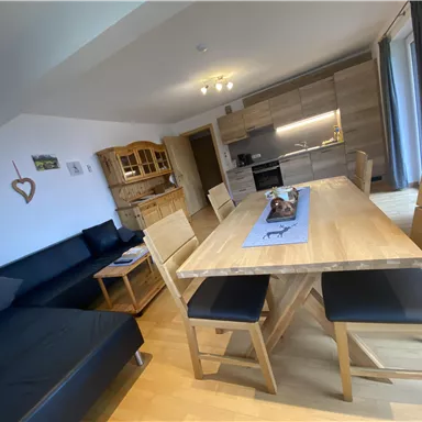 A modern living room with a bright kitchen. There is a large wooden table with chairs and a comfortable couch.