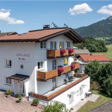A modern building with balconies, surrounded by green meadows and mountains. The facade is bright and friendly, with colorful flower boxes.