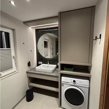 A modern bathroom with a round mirror and a washing machine. The space is bright and functional with a simple design.