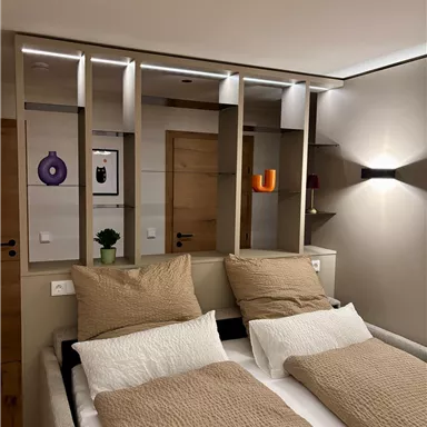 A modern bedroom with two single beds and neutral colors. In the background, there are decorative shelves and artwork on the wall.