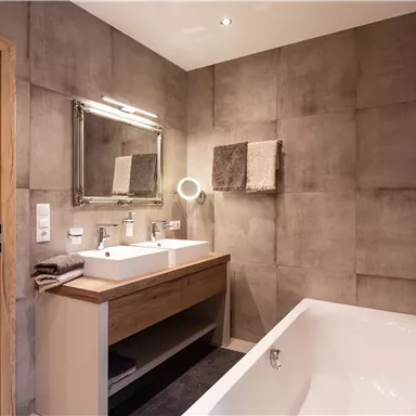 A modern bathroom with an elegant bathtub and two sinks. The walls are in a neutral gray tone, complemented by warm wooden accents.
