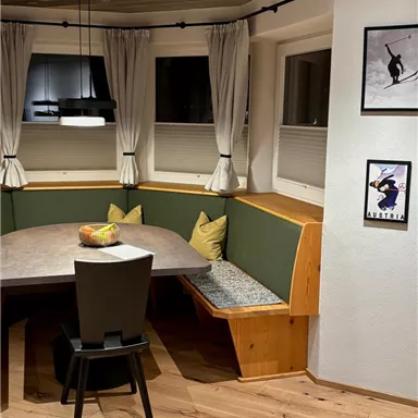 A cozy dining area with a corner bench, a large table, and modern chairs. The walls are decorated with pictures and light curtains.