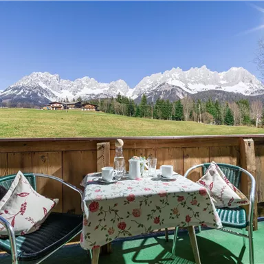 Eine gemütliche Terrasse mit einem Tisch und Stühlen, umgeben von einer schönen Berglandschaft. Im Hintergrund sind schneebedeckte Berge und ein klarer blauer Himmel zu sehen.