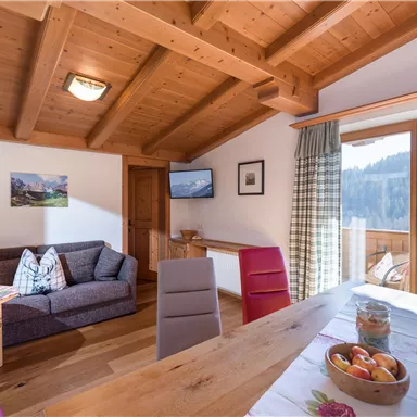 Ein gemütliches Wohnzimmer mit Holzbalkendecke und einem Esstisch. Große Fenster bieten einen Blick auf die umliegende Natur.