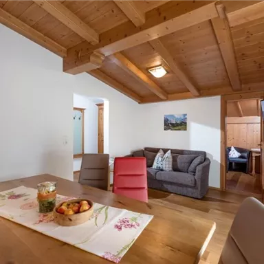 Eine helle und gemütliche Wohnküche mit Holzdecke und einem großen Esstisch. Im Hintergrund sieht man eine Sitzecke und einen Fernseher.