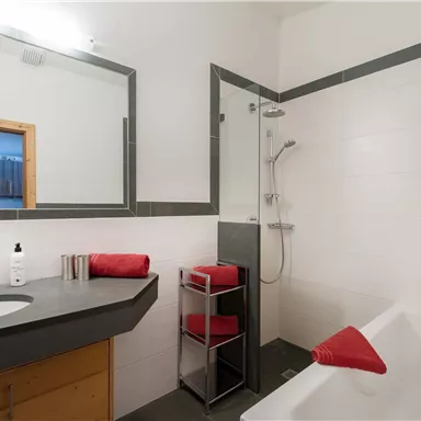 Ein modernes Badezimmer mit einer Dusche, Badewanne und einem großen Spiegel. Rote Handtücher und Pflegeprodukte sorgen für einen einladenden Eindruck.