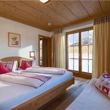 Ein gemütliches Schlafzimmer mit zwei Betten und Holzmöbeln. Große Fenster sorgen für viel Tageslicht und bieten einen Blick nach draußen.