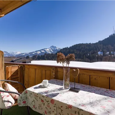 Ein gemütlicher Balkon mit einem Tisch und Stühlen. Im Hintergrund erstreckt sich eine wunderschöne Berglandschaft unter klarem blauem Himmel.
