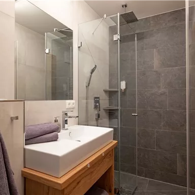 Ein modernes Badezimmer mit einer walk-in Dusche und einem Waschbecken. Die Wände sind in neutralen Farben gehalten und der Boden ist mit grauen Fliesen ausgelegt.