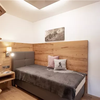 Ein gemütliches Zimmer mit Holzverkleidung und einem Einzelbett. An der Wand hängt ein Fernseher, und ein kleines Fenster bietet Blick auf die Berge.