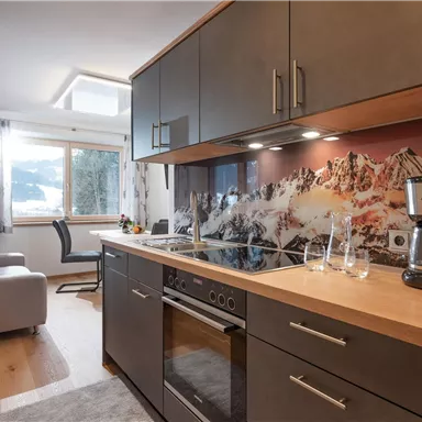 Eine moderne Küche mit einem Holzarbeitsplatz und eleganten Schränken. Im Hintergrund ist ein gemütlicher Wohnbereich mit einem Fenster und einem schönen Bergmotiv zu sehen.