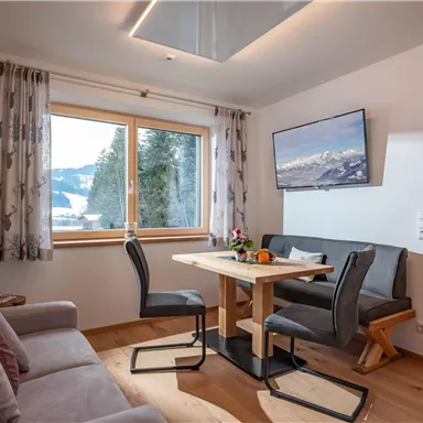 Ein gemütlicher Wohnraum mit einer Sitzgruppe und einem Esstisch. Große Fenster bieten einen Blick auf die Berge und sorgen für viel Tageslicht.