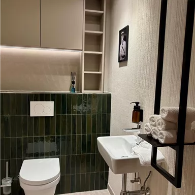 A modern bathroom with green tiles and an elegant sink. On the wall, there are towels and toiletries as well as a decorative picture.