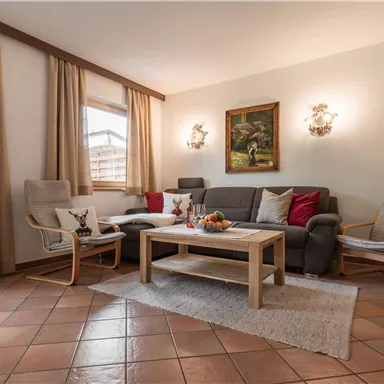 Ein gemütliches Wohnzimmer mit einer grauen Couch und zwei beigen Sesseln. Der Tisch ist mit Obst und einer Blumenvasen dekoriert, und es gibt große Fenster mit Vorhängen.