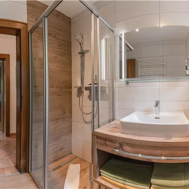 Ein modernes Badezimmer mit einer Dusche und einem Holzwaschtisch. Die Wände sind hell gefliest, und es gibt einen großen Spiegel mit Beleuchtung.